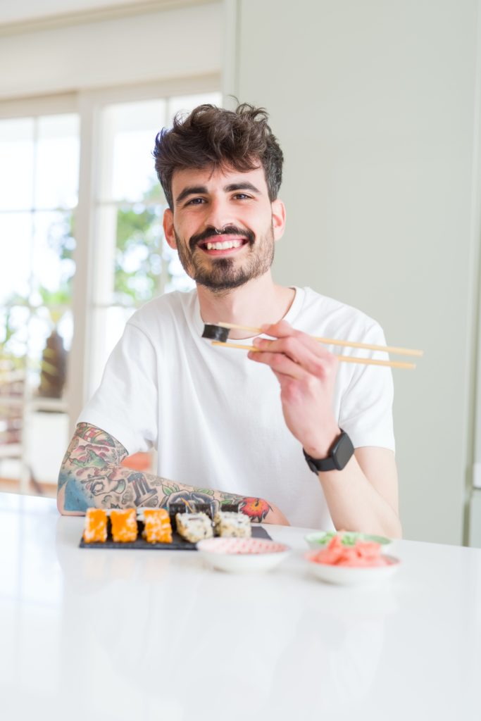 hombre sentado con sushi de salmón al frente, sosteniendo con palitos un rollo de alga